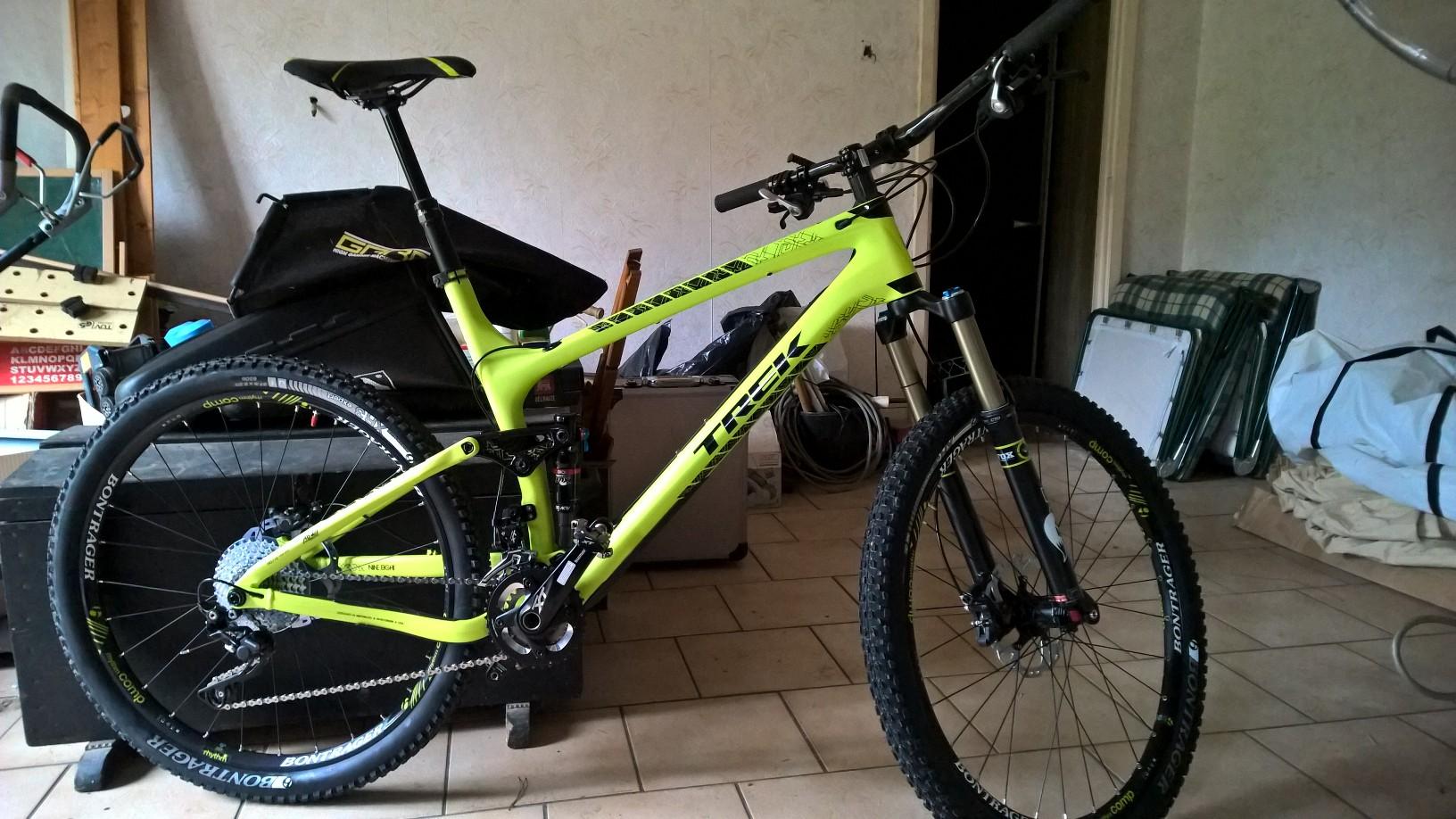 Trek Trek Fuel EX 9.8 27.5: A bright green Trek mountain bike is positioned in a cluttered indoor space, showcasing its sturdy frame, front suspension, and knobby tires. The background contains various items, including boxes and a folded chair, adding to the casual, home environment.