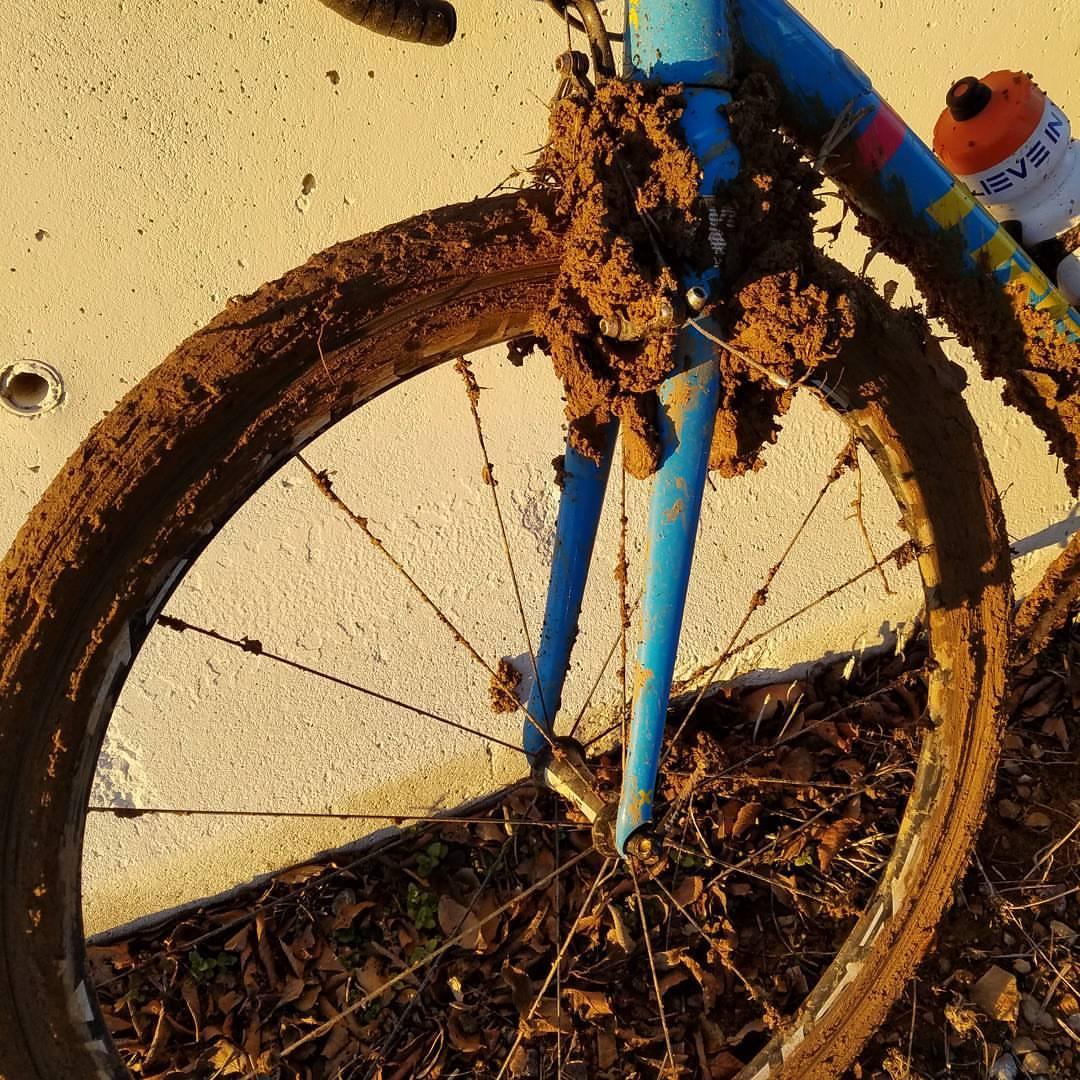 Close-up of a muddy bicycle leaning against a textured white wall. The bike features a blue frame with dirt caked on the front wheel, tires, and fork, indicating it has been used in muddy conditions. Fallen leaves are scattered on the ground beneath the bike. Valley Trail mountain bike trail.
