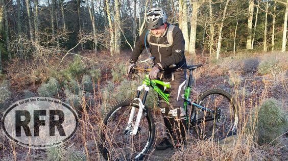 A mountain biker in protective gear is standing next to a green and black bike on a rugged trail surrounded by trees and tall grass. The sun is shining, indicating a clear day. The biker is focused on adjusting something on the bike while wearing a helmet and cycling attire. Village Park/ Abrams Rock mountain bike trail.