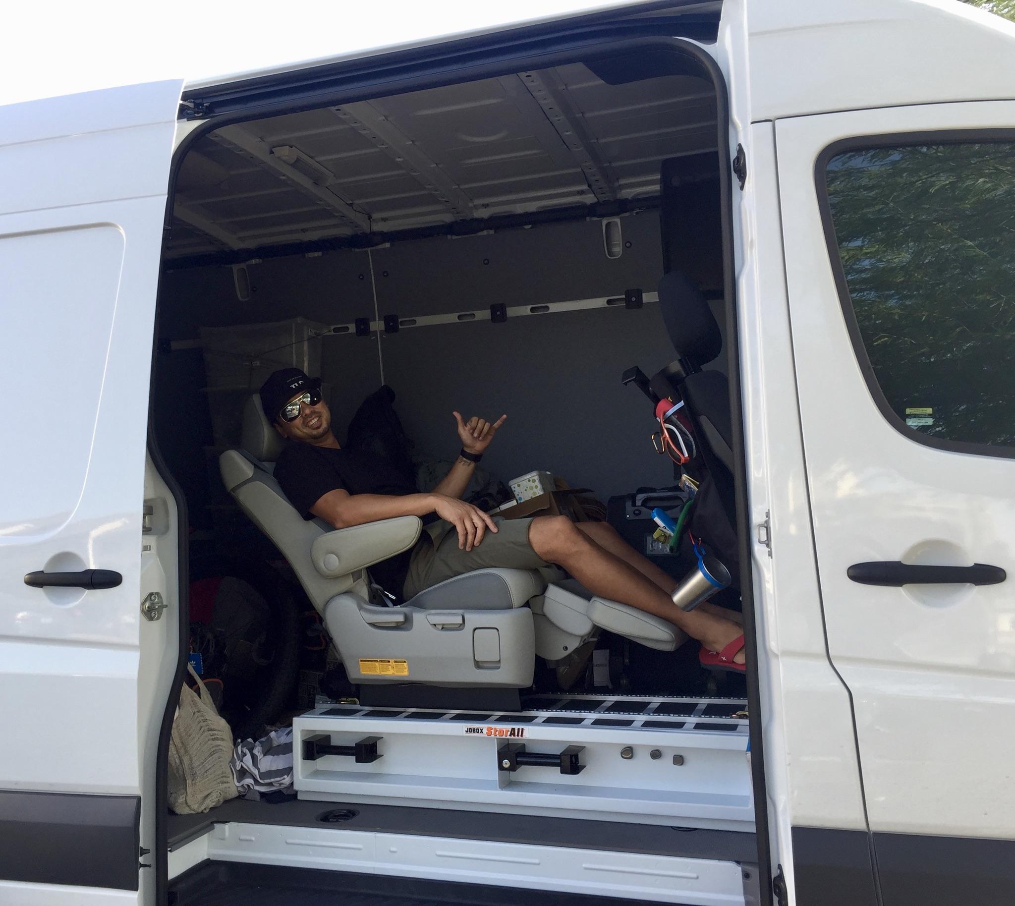 Specialized Stumpjumper: A person sitting in the driver's seat of a van, smiling and making a hand gesture, with the interior of the van partially visible, showing storage items and an organized space.