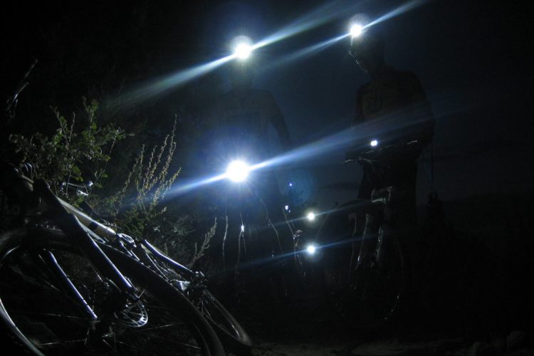 Two cyclists stand in the dark wearing headlamps that illuminate their surroundings. A bicycle lies on the ground in the foreground, while sparse vegetation is visible in the background. The bright beams of light from the headlamps create an atmospheric, nighttime scene.
