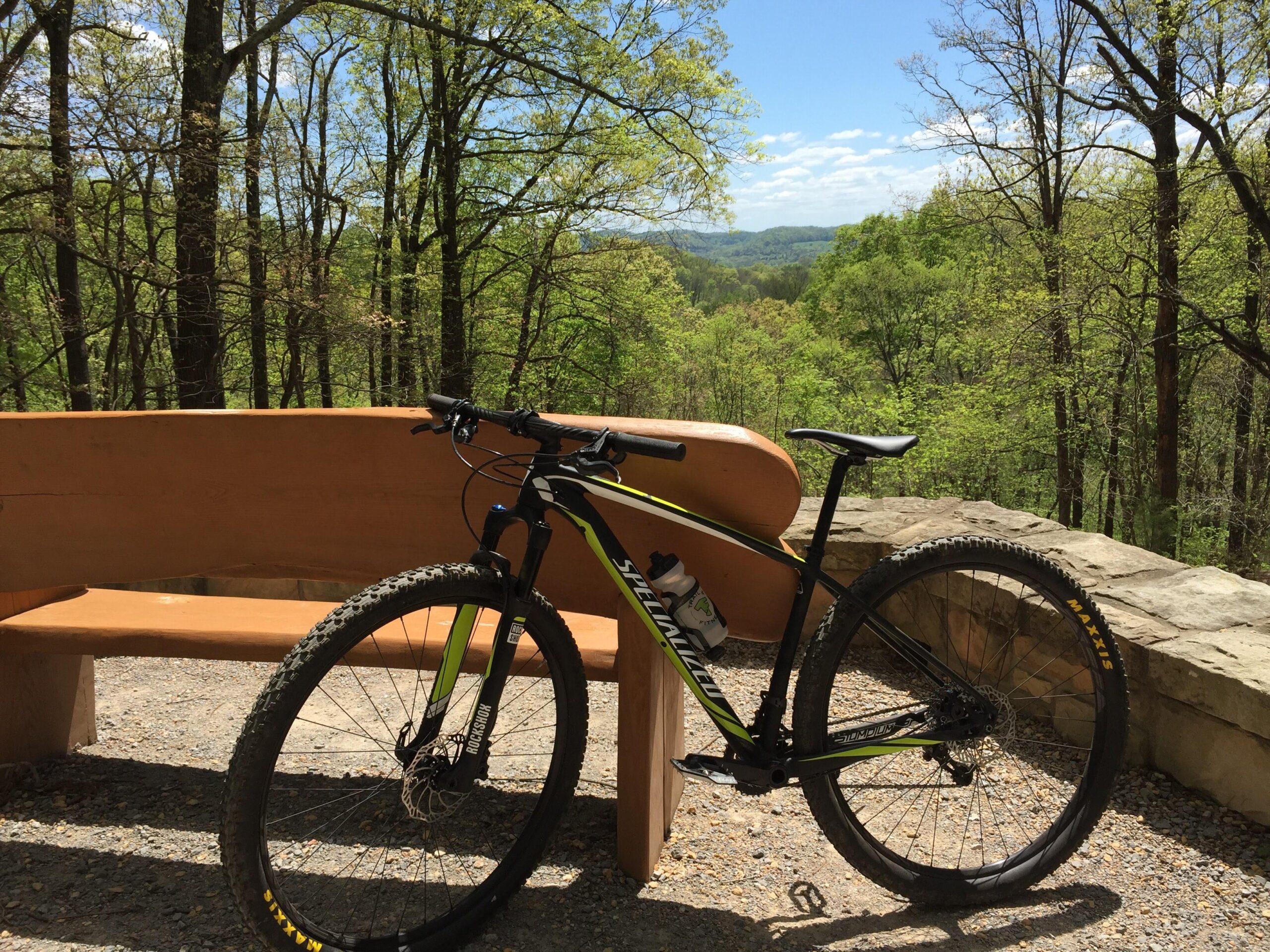 Specialized Stumpjumper Comp 29er: A mountain bike resting beside a wooden bench, with a scenic view of a lush green forest and rolling hills in the background under a clear blue sky.