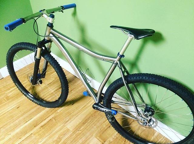 Carver 420: A mountain bike with a silver frame and thick tires, leaning against a green wall. The bike features blue handlebars and a black seat, with visible suspension in the front fork. The floor is wooden, and the setting appears to be indoors.