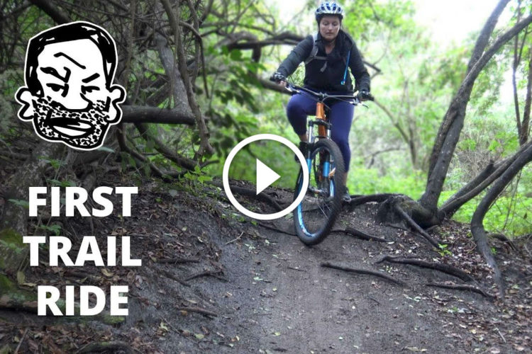 A person riding a mountain bike on a dirt trail surrounded by trees and foliage, navigating a section with roots and uneven terrain. The image includes a graphic overlay that says "FIRST TRAIL RIDE."