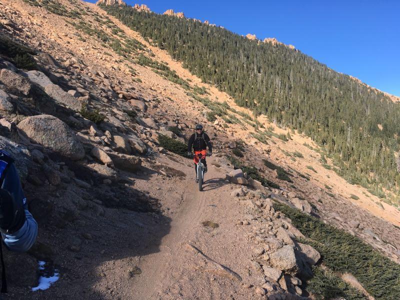 A cyclist riding along a rocky mountain trail surrounded by sparse vegetation and pine trees under a clear blue sky. The path is narrow and steep, leading through a rugged landscape with scattered rocks. Elk Park Trail mountain bike trail.