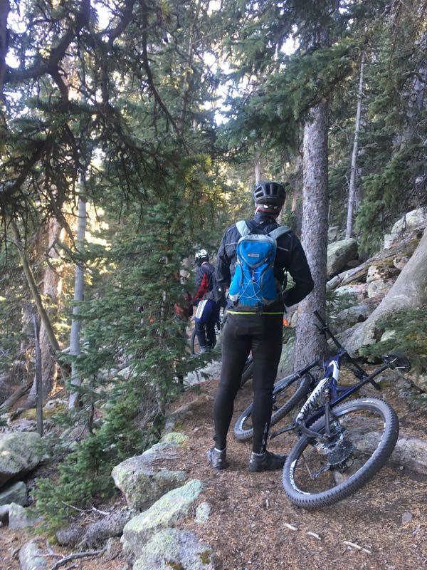 A mountain biker stands on a narrow trail surrounded by tall trees and rocky terrain, looking ahead. A bicycle is parked beside them, and another cyclist can be seen in the background. The scene captures the natural beauty of the forest and the adventure of mountain biking. Elk Park Trail mountain bike trail.