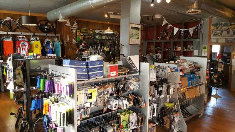 An indoor view of a bicycle shop showcasing a variety of cycling gear and accessories. The store features clothing hanging on the walls, organized displays of bike parts and tools, and a well-lit atmosphere. Shelves are stocked with colorful items, including grips and accessories, while bicycles and cycling-related items are visible throughout the space.
