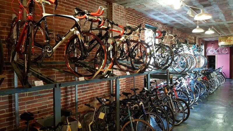 A bike shop interior showcasing various bicycles. Some bikes are displayed on shelves with a brick wall backdrop, while many others are lined up on the floor. The lighting is bright, highlighting the different models and colors of the bikes.