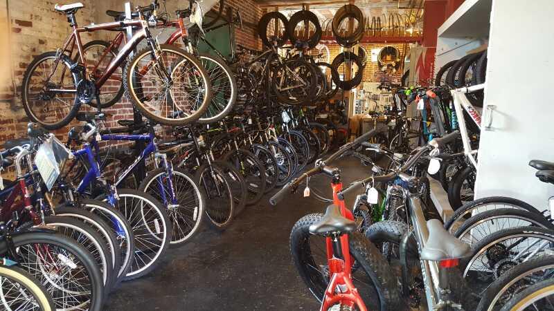 A bike shop interior displaying a variety of bicycles lined up on the floor and hung on walls, with tires and bike accessories visible in the background. The room features exposed brick walls, creating an industrial atmosphere.