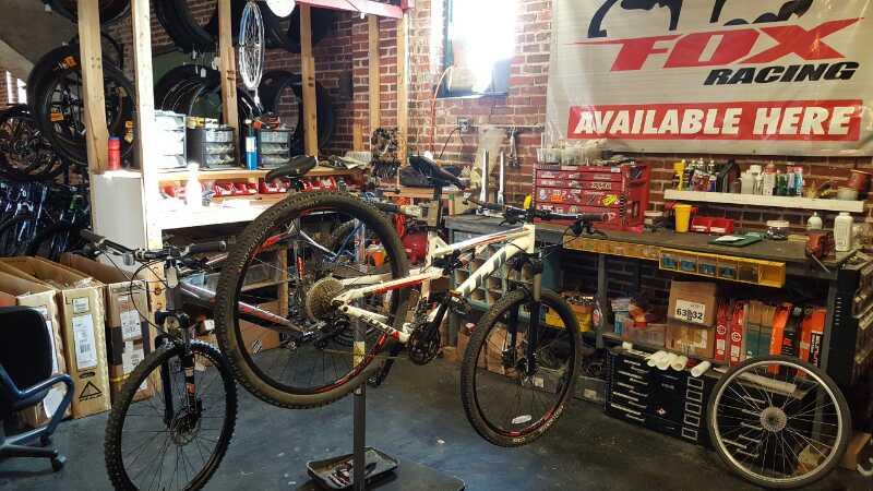 A bike repair shop interior featuring an elevated mountain bike on a stand, surrounded by various bike parts and tools. The background includes shelves with bike wheels and a workbench with repair supplies. A sign reading "FOX Racing Available Here" is prominently displayed on the wall.
