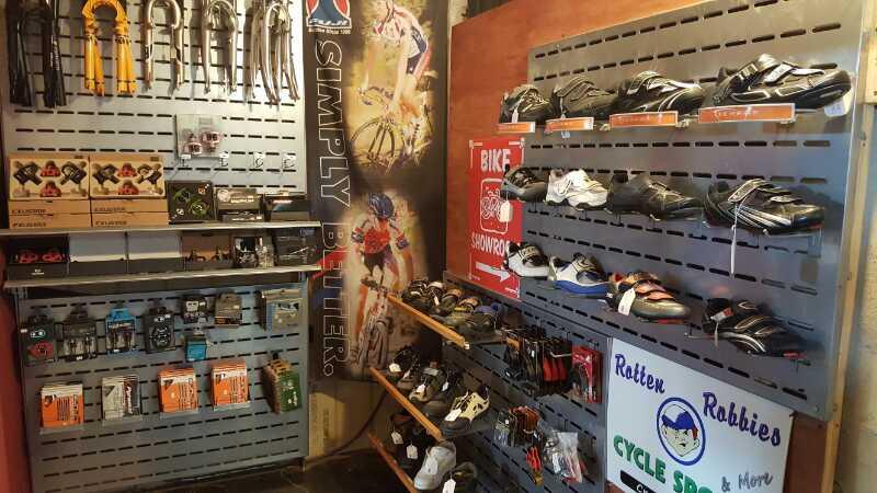 A display in a bike shop showcasing various cycling accessories and shoes. The left wall features a range of bike parts and tools organized on pegboards, while the right side displays several pairs of cycling shoes. Promotional banners and a sign for "Rotten Rabbies Cycle Shop" are visible in the background.