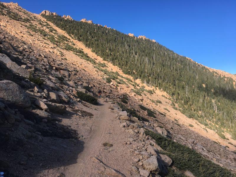 A winding dirt trail cuts through rocky terrain, leading up a slope covered with sparse vegetation and tall evergreen trees in the background. The scene is set under a clear blue sky, giving a sense of serenity and adventure in a mountainous landscape. Elk Park Trail mountain bike trail.