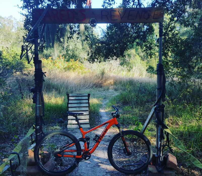 A mountain bike in front of a wooden archway that reads "Jared's Revenge." The path leads into a grassy area surrounded by trees. The archway is supported by wooden and metal posts, with additional biking gear visible nearby. Loyce E. Harpe Park mountain bike trail.