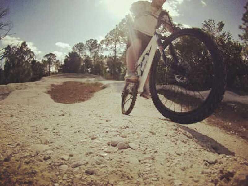 A cyclist performing a trick by lifting the front wheel of a mountain bike on a dirt trail surrounded by trees, with sunlight shining down from above. Jonathan Dickinson State Park mountain bike trail.