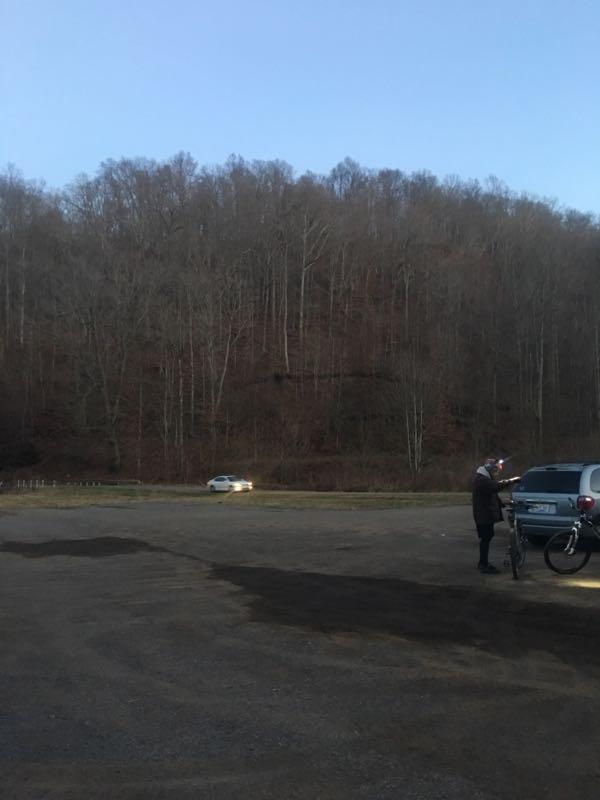 A person standing next to a bicycle, looking at a phone, in a gravel parking area. In the background, a wooded hillside with bare trees is visible under a clear sky. A car is driving away on a nearby road. Youngers Creek mountain bike trail.
