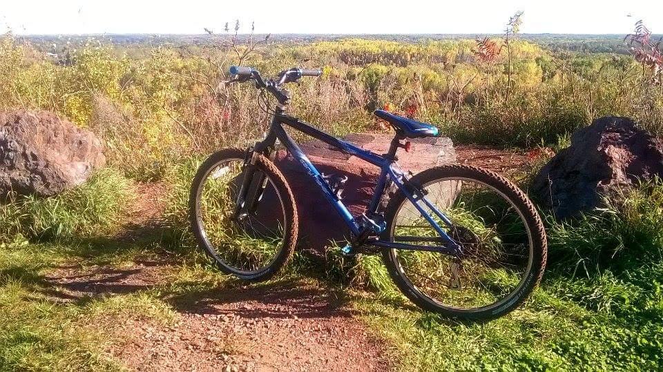 A mountain bike leaning against a large rock, set on a dirt path surrounded by lush green grass and wildflowers. In the background, a scenic view of rolling hills and a clear sky. The scene is bright and sunny, suggesting an ideal day for outdoor activities. Cuyuna Lakes mountain bike trail.