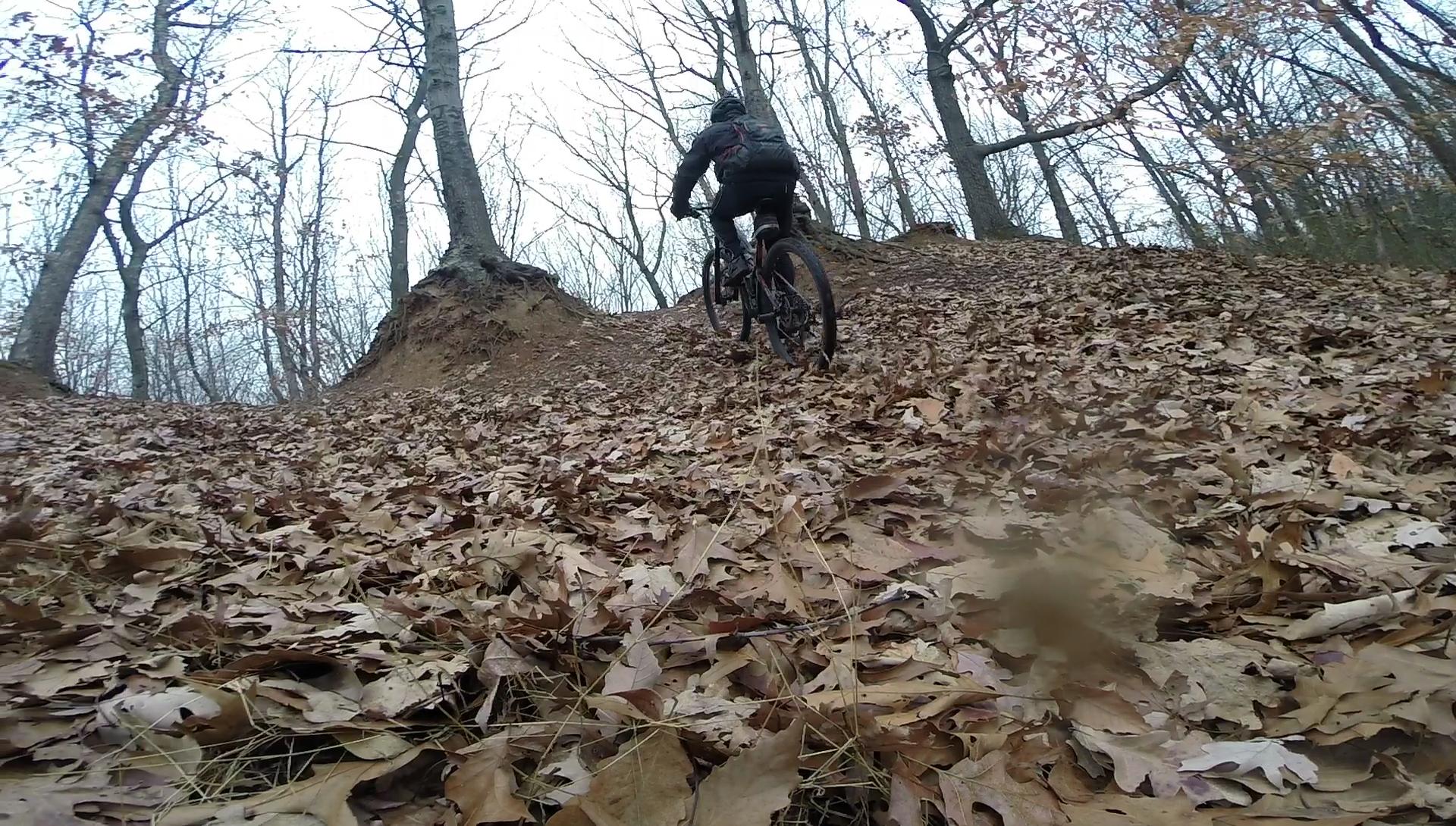 A mountain biker ascends a leaf-covered trail in a wooded area during autumn, surrounded by trees with bare branches. The ground is scattered with fallen leaves, and the trail features a slight incline leading upwards. Long Pond mountain bike trail.