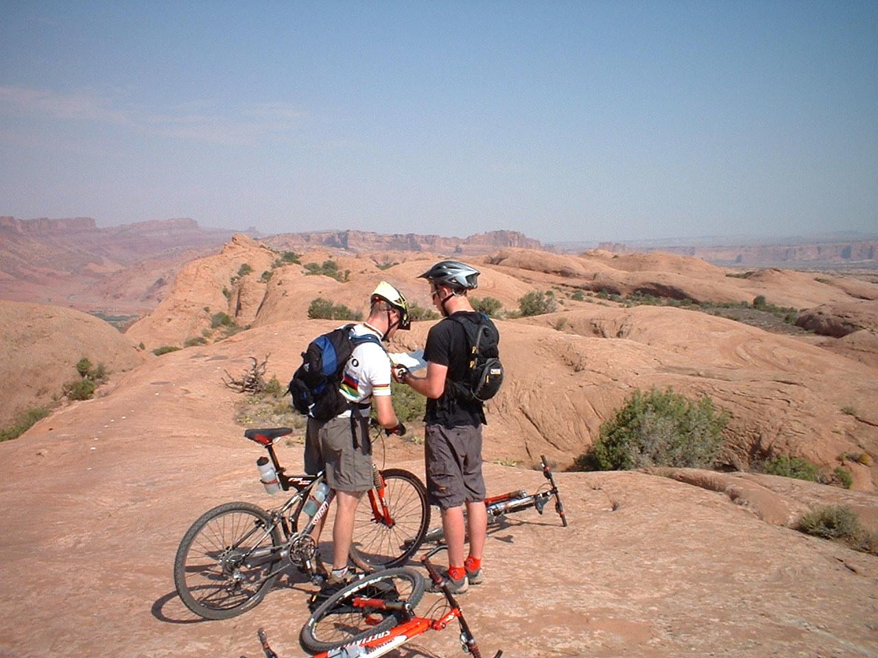 Gary Fisher Hoo Koo E Koo: Two mountain bikers standing on a rocky terrain, checking a map or GPS device, with bikes nearby. The landscape features undulating red rock formations and distant mountains under a clear blue sky.