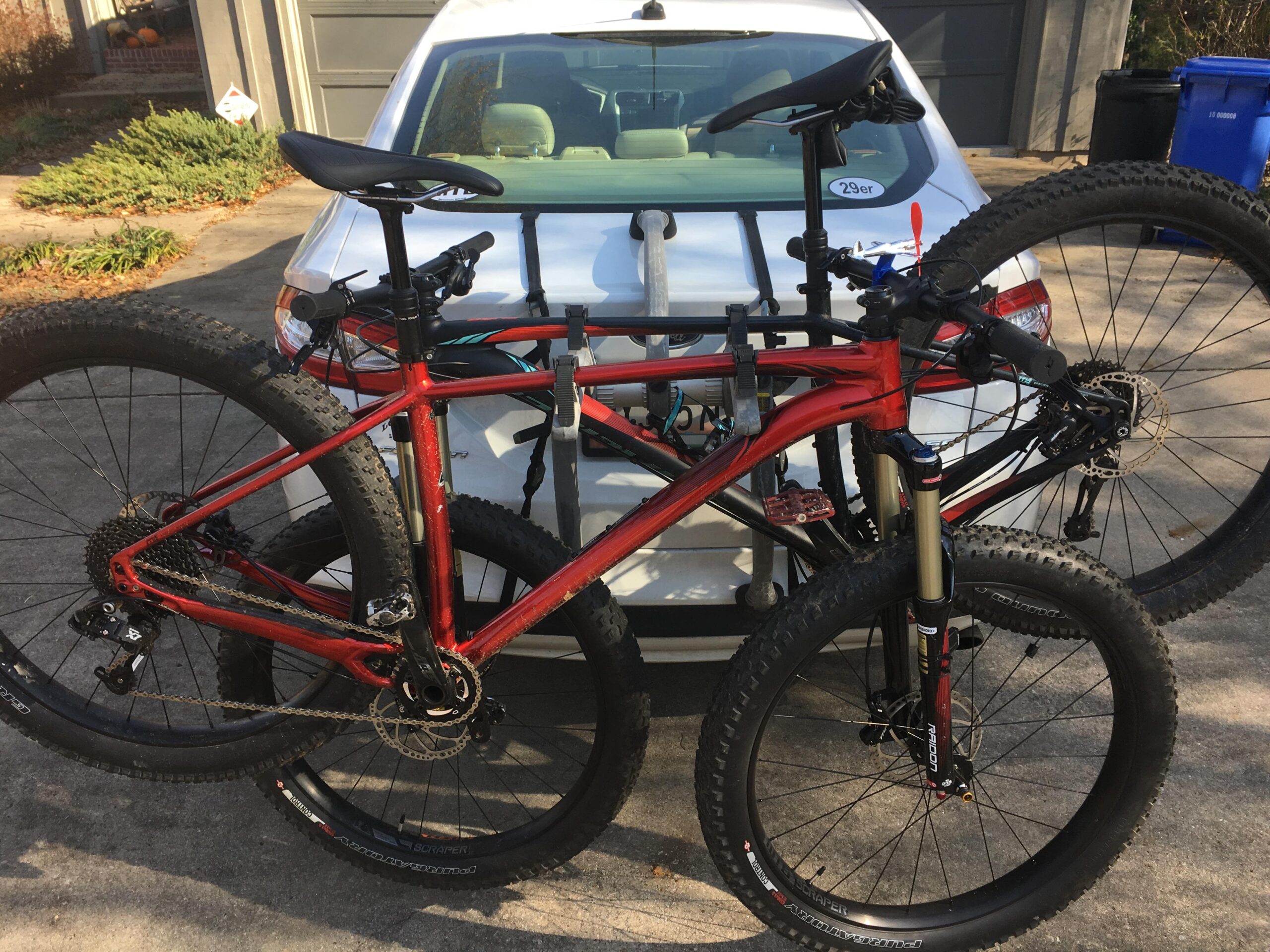 Specialized Fuse Comp 6Fattie: A red mountain bike secured on a bike rack attached to the back of a white car. The bike has large, knobby tires and a modern design, with various gears and components visible. The background includes a glimpse of a house and some greenery.