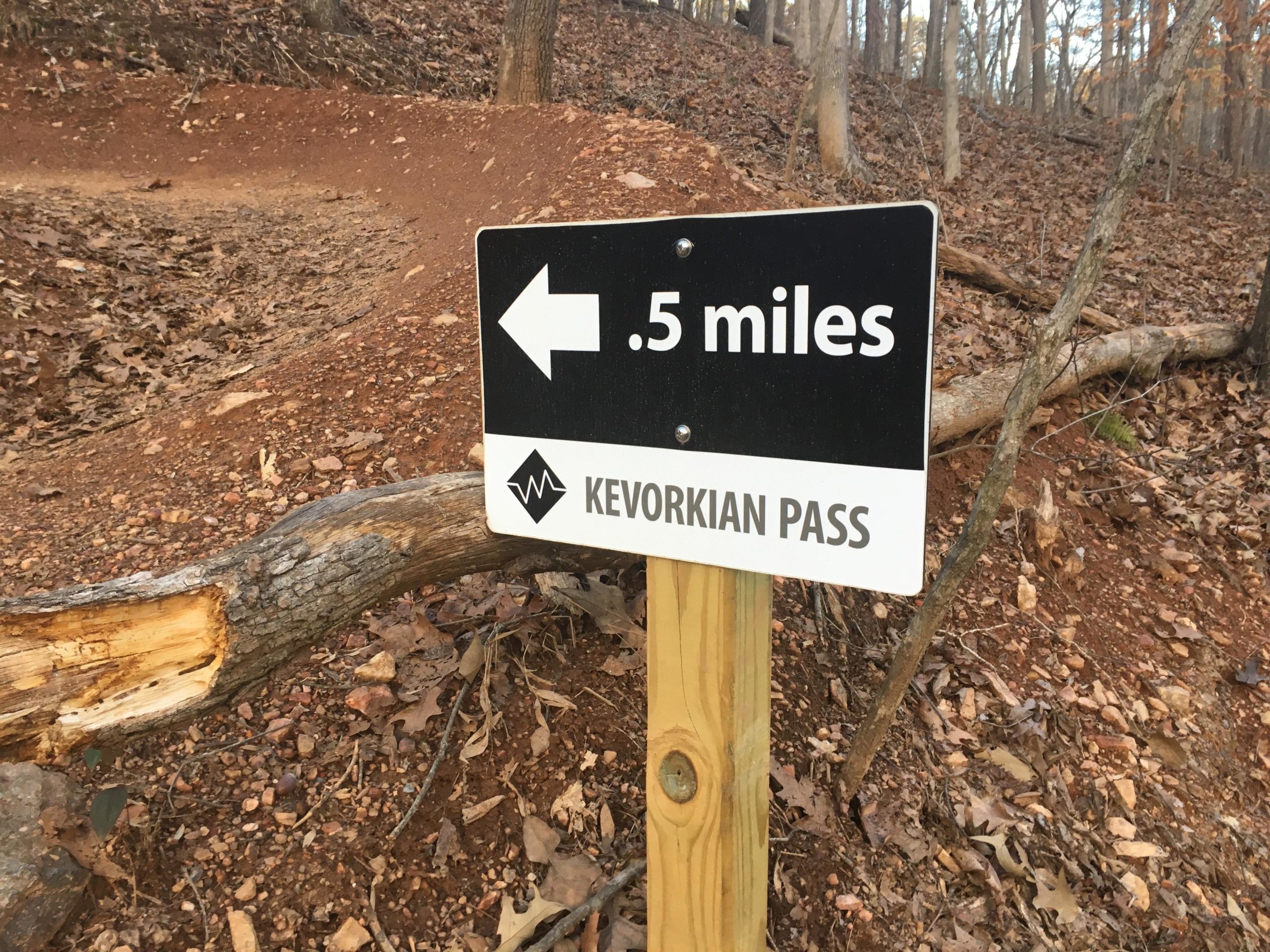 A trail sign indicating "Kervorkian Pass" with an arrow pointing left and a distance of 0.5 miles. The sign is mounted on a wooden post in a wooded area with fallen leaves on the ground. Van Michael Trail mountain bike trail.