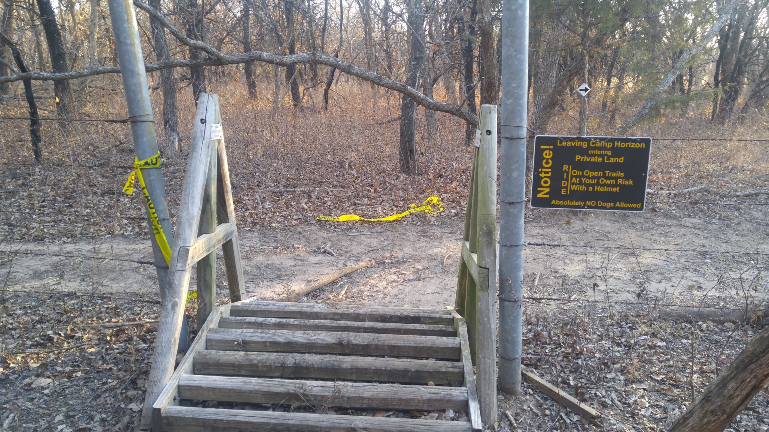 A wooden staircase leading into a wooded area, marked by a sign that reads "Leaving Camp Horizon, entering Private Land" with additional warnings about riding on open trails at one’s own risk and a note that no dogs are allowed. Bright yellow caution tape is visible on the ground and near a metal post. The surrounding area features bare trees and dried leaves. Camp Horizon mountain bike trail.
