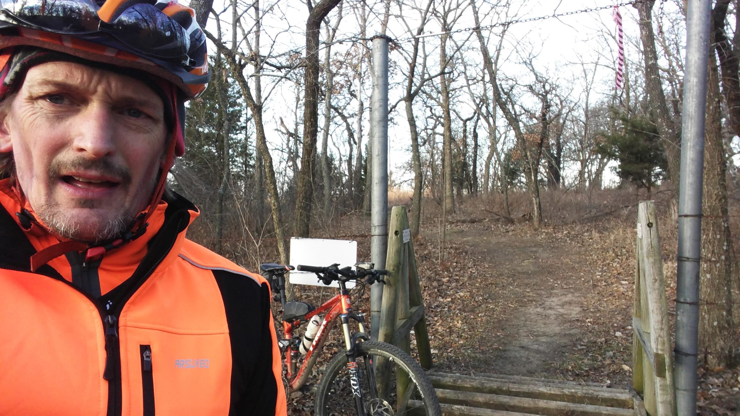 A cyclist wearing a bright orange jacket and helmet stands near a mountain bike, positioned by a wooden bridge that leads into a wooded trail. The background features bare trees and a clear sky, indicative of a cool day. Camp Horizon mountain bike trail.
