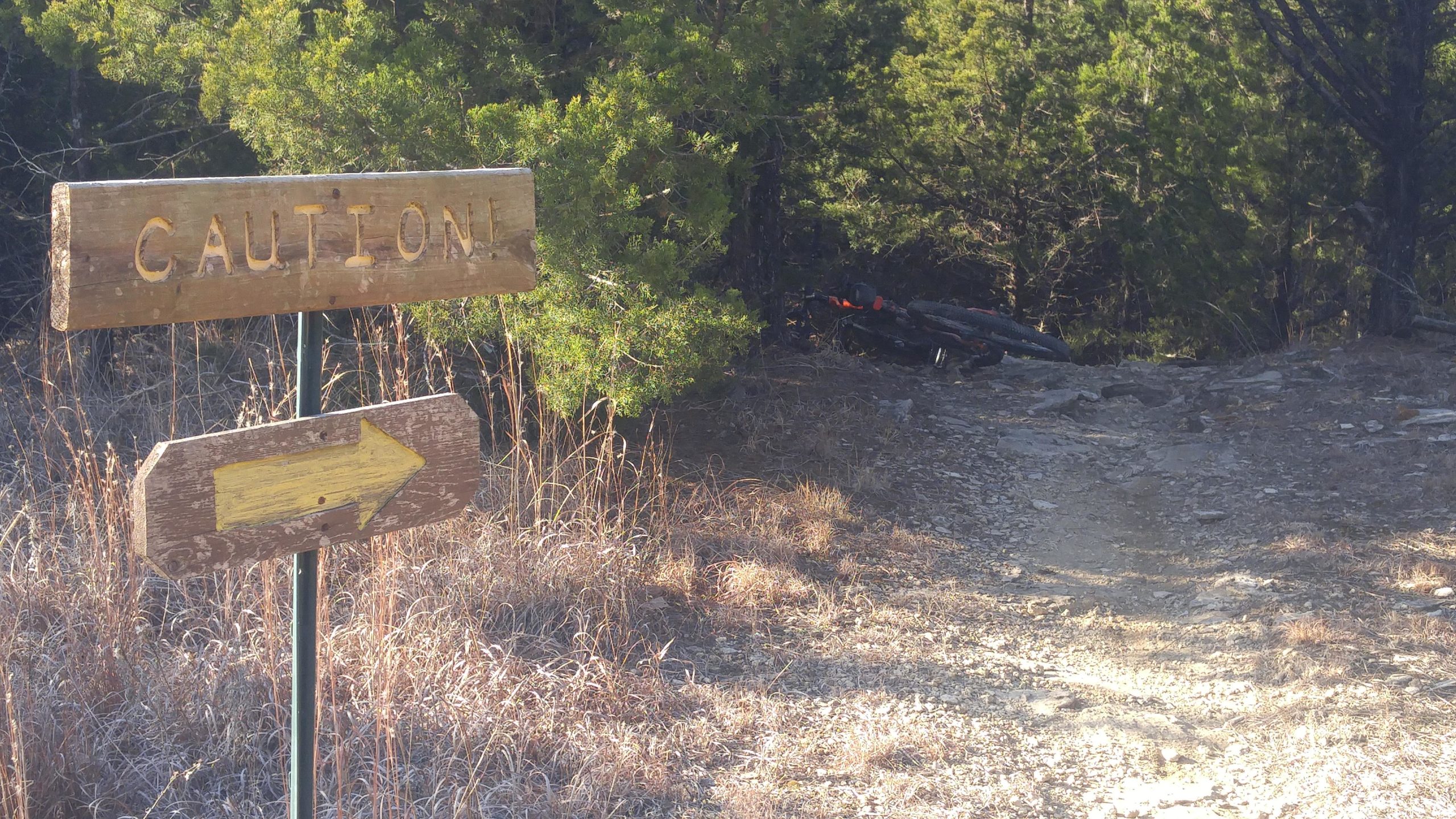 A wooden sign with the word "CAUTION!" prominently displayed, pointing to the right with an arrow. The background features a narrow, rocky path surrounded by vegetation, with a hint of a bicycle partially obscured by trees. Sunlight casts a warm glow on the scene, highlighting the overgrown grass and rugged terrain. Camp Horizon mountain bike trail.