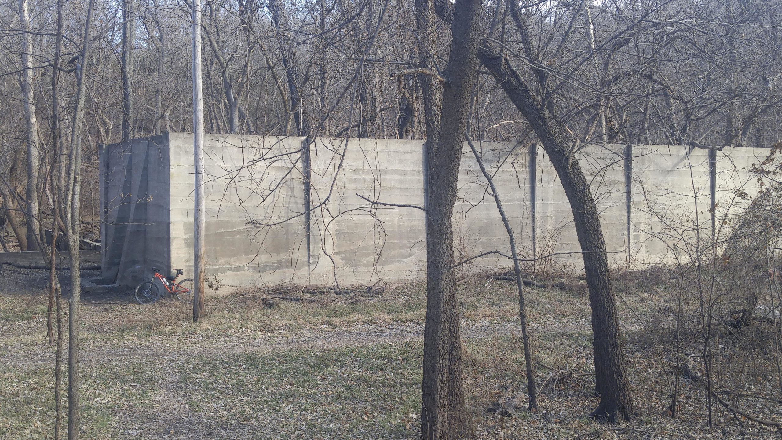 A concrete wall stands in a wooded area during the winter, with bare trees and a patch of grass in the foreground. An orange bicycle is leaning against the wall, and a gravel path can be seen leading through the scene. Camp Horizon mountain bike trail.