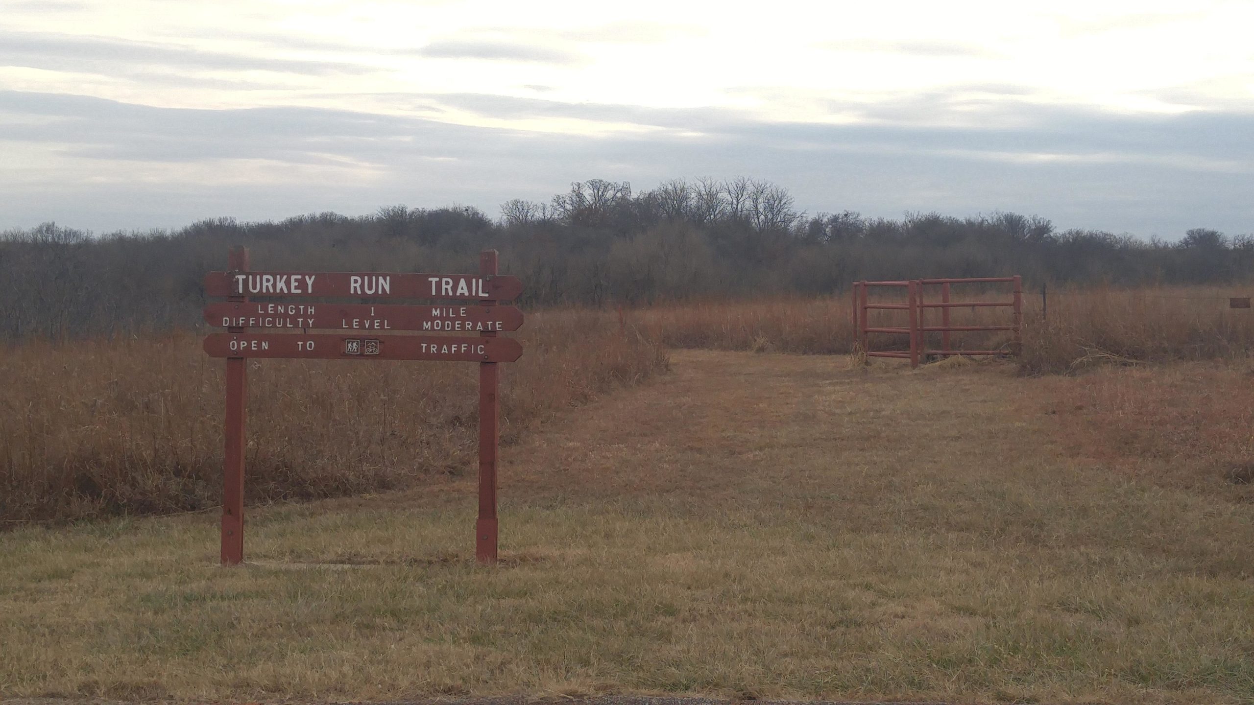 Sign for Turkey Run Trail indicating a length of 1 mile, moderate difficulty level, and open to traffic, set against a backdrop of tall grass and trees under a cloudy sky. Turkey Run Trail mountain bike trail.