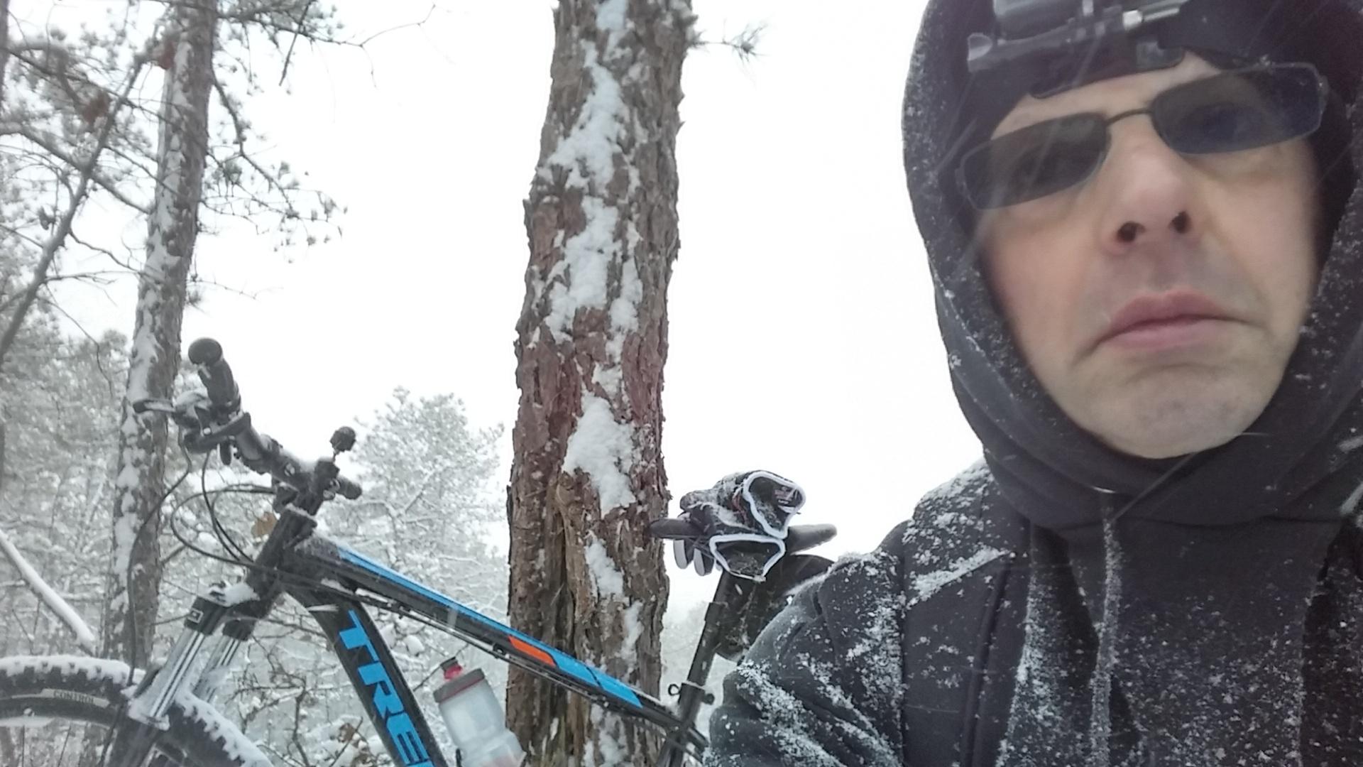 A person in winter clothing stands in a snowy forest, capturing a selfie with a mountain bike beside them. The bike has snow accumulating on its frame, and snow covers the ground and nearby trees. The individual wears sunglasses and a head-mounted camera, with snowflakes visible in the air. Forked River Mountain mountain bike trail.