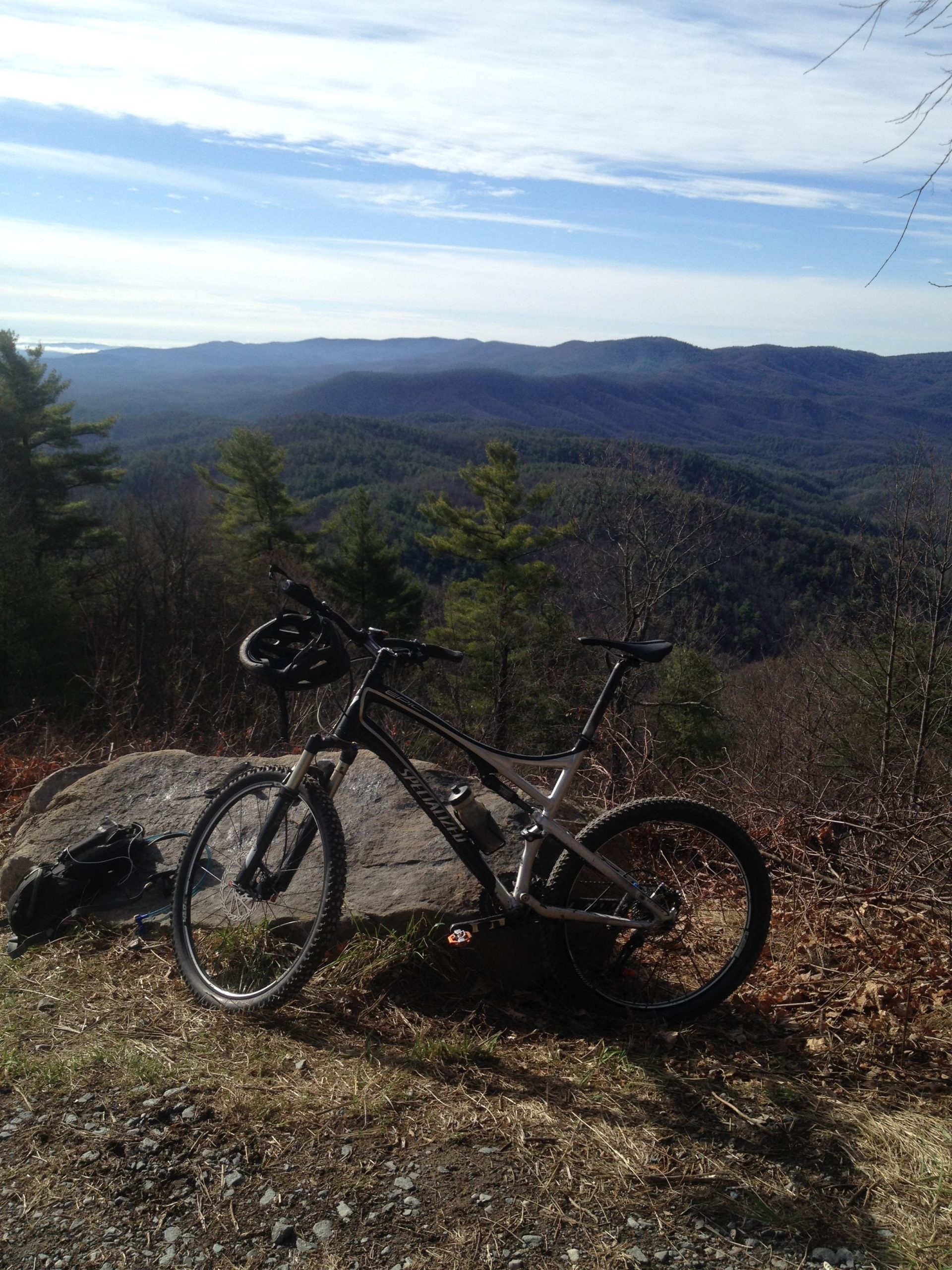 A mountain bike rests against a large rock, with a scenic view of rolling mountains and a clear blue sky in the background. Pine trees dot the landscape, showcasing a tranquil outdoor setting suitable for biking and exploring. Pinhoti Trail: P1 / Bear Creek Pinhoti mountain bike trail.