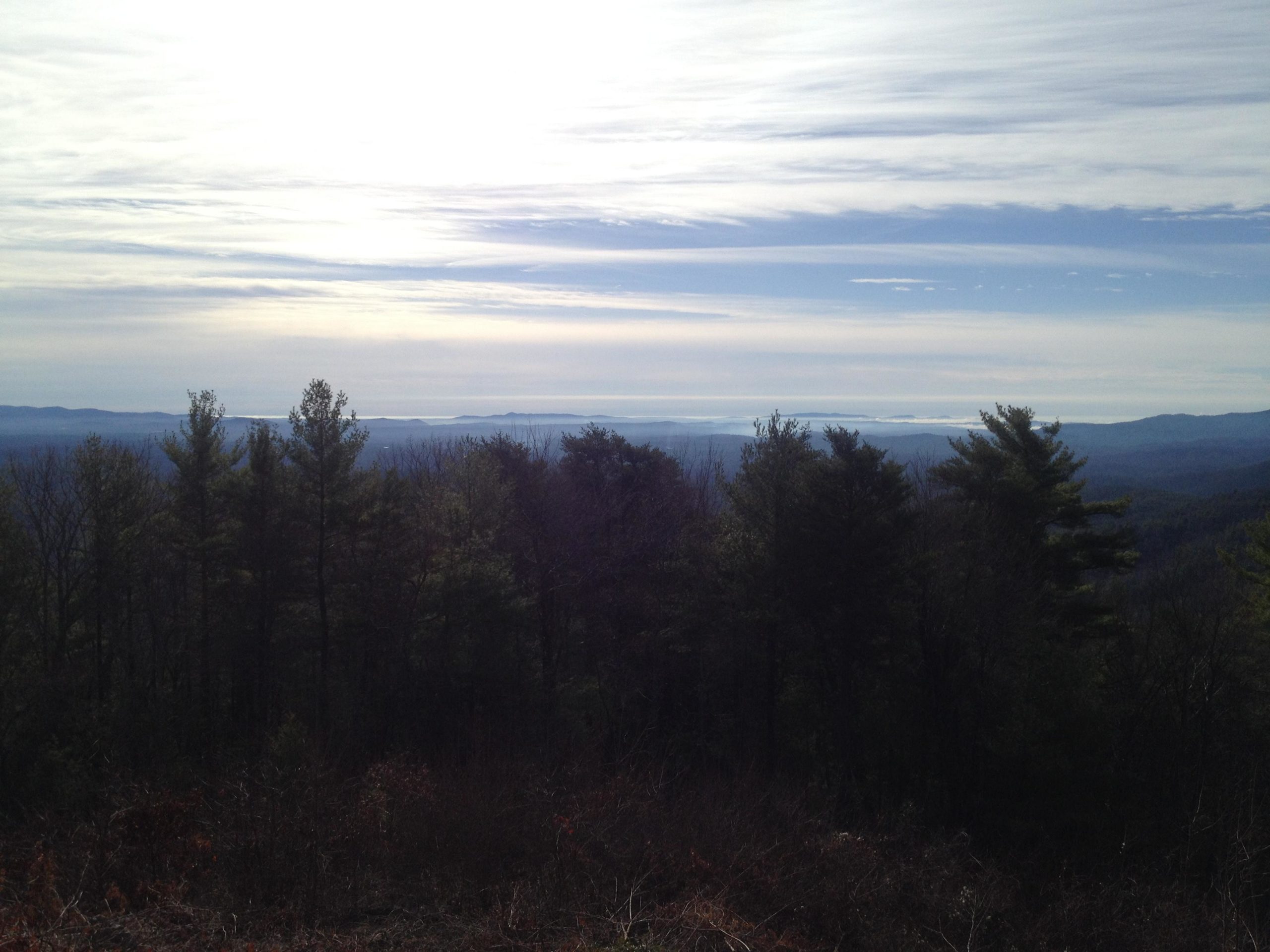 A panoramic view of a mountainous landscape with a forested foreground. The sky is partly cloudy, illuminated by soft sunlight creating a serene atmosphere. In the distance, rolling hills and mountains are visible under a hazy horizon, suggesting depth and natural beauty. Pinhoti Trail: P1 / Bear Creek Pinhoti mountain bike trail.