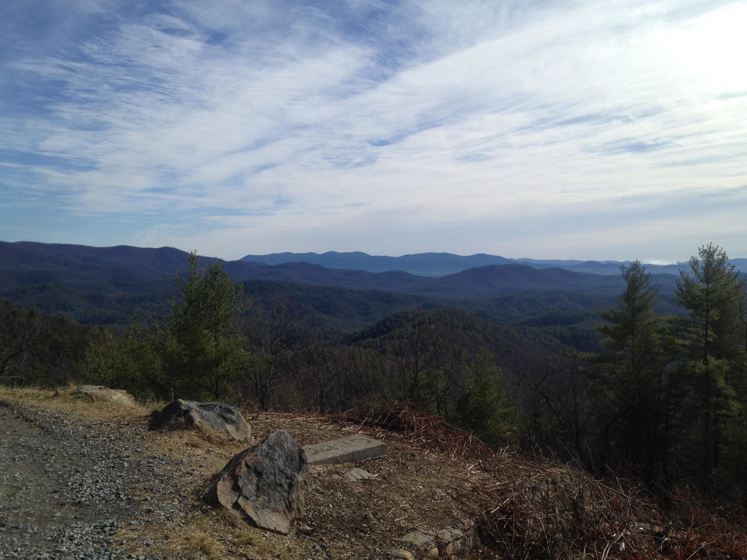 A panoramic view of mountainous landscape under a partly cloudy sky, showcasing layers of rolling hills and valleys covered with lush greenery and scattered trees. The foreground features rocky outcrops and a gravel path, enhancing the natural scenery. Pinhoti Trail: P1 / Bear Creek Pinhoti mountain bike trail.