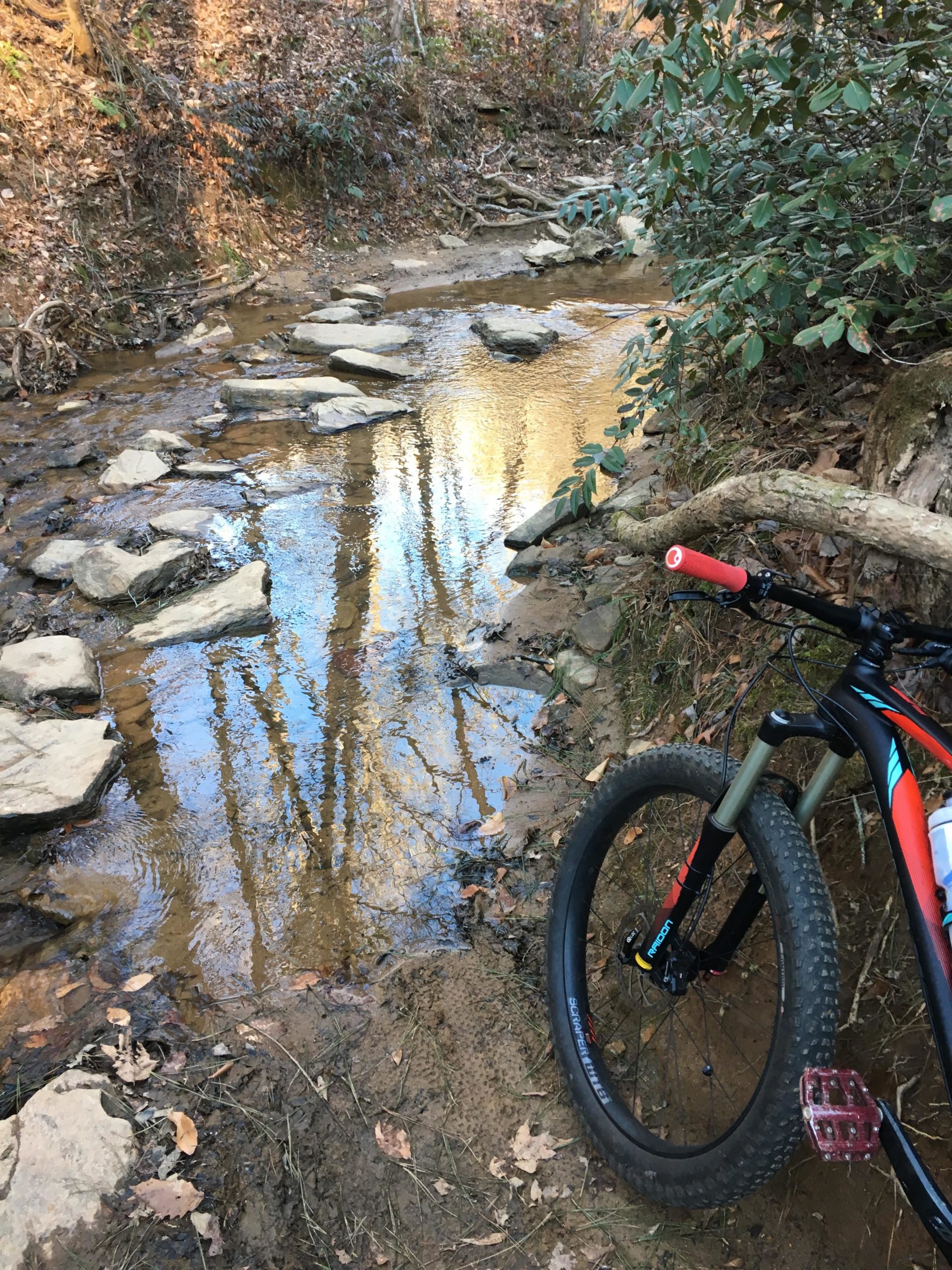 A mountain bike rests on the bank of a shallow creek, with large flat stones forming a natural crossing over the water. Surrounding the creek is a forested area with trees and fallen leaves, reflecting the warm tones of the setting sun. The scene captures a peaceful outdoor setting ideal for biking and exploration. Chicopee Woods mountain bike trail.