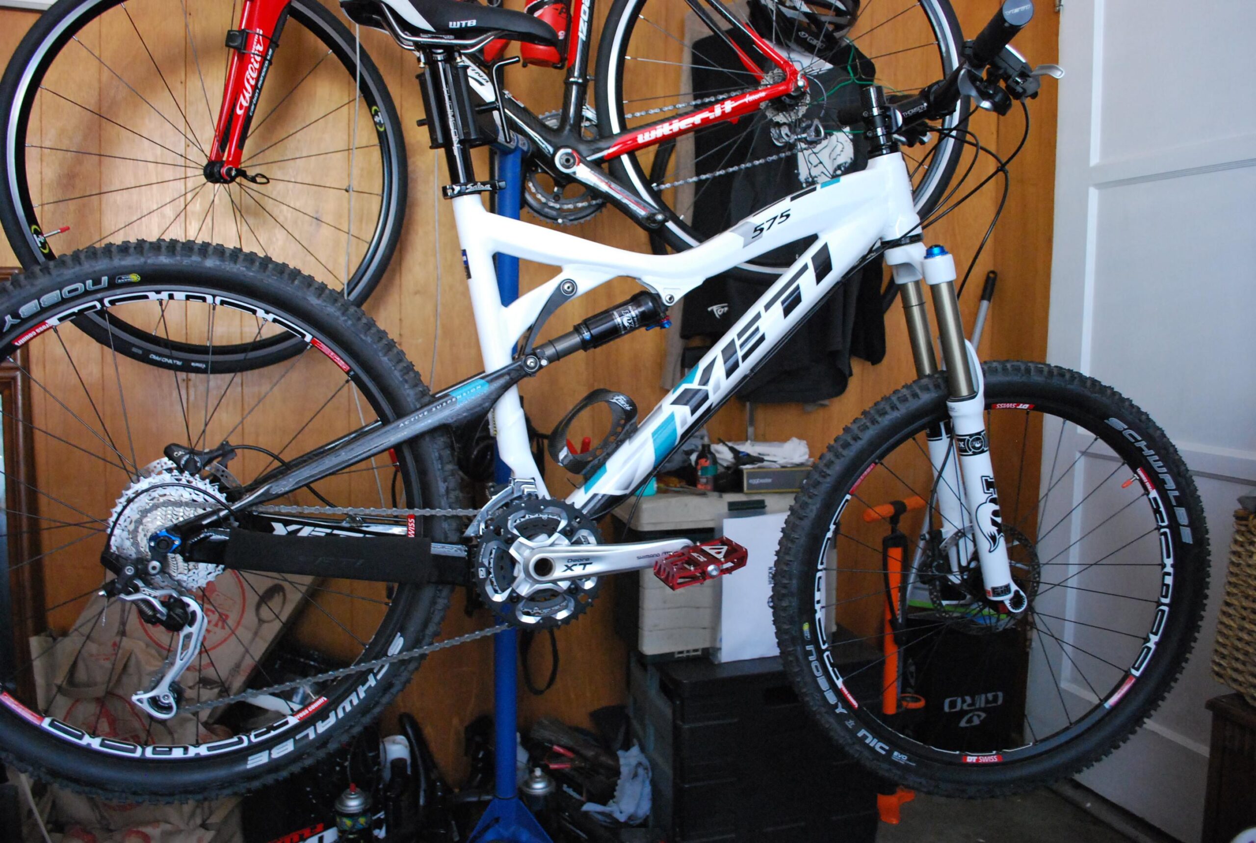 Yeti 575: A close-up image of a white Yeti mountain bike hanging in a garage. The bike features a prominent suspension system, Shimano XT drivetrain, and red pedals. In the background, another bike can be seen along with various tools and equipment typical of a biking workshop.