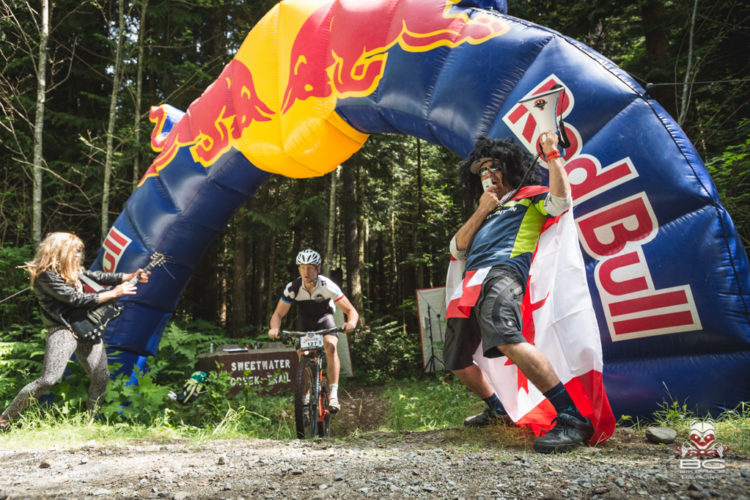 A mountain biker rides through a colorful inflatable archway marked with the Red Bull logo. To the left, a musician plays an electric guitar, while on the right, a person dressed in a colorful outfit and a wig waves a flag and cheers enthusiastically. The scene is set in a forested area, capturing the excitement of a cycling event.