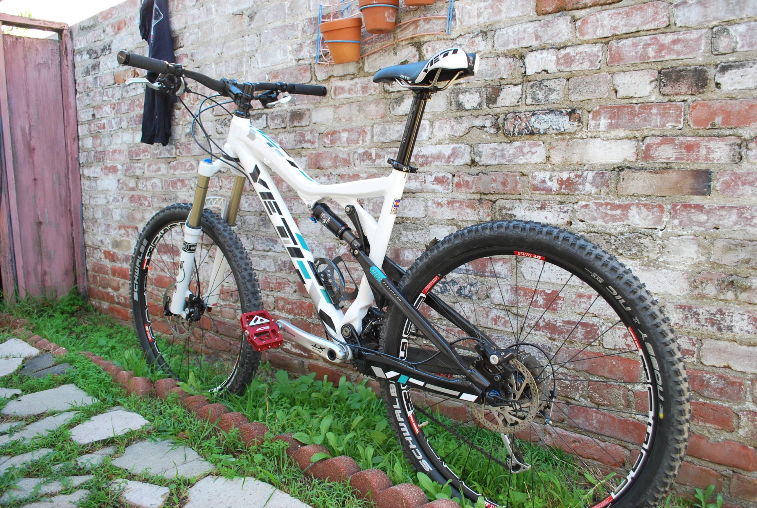 Yeti 575: A white mountain bike leaning against a brick wall, featuring black and blue accents, with a visible red pedal. The scene includes patches of grass and a brick path. A black shirt is hanging on the wall, and there are small potted plants mounted on the wall.