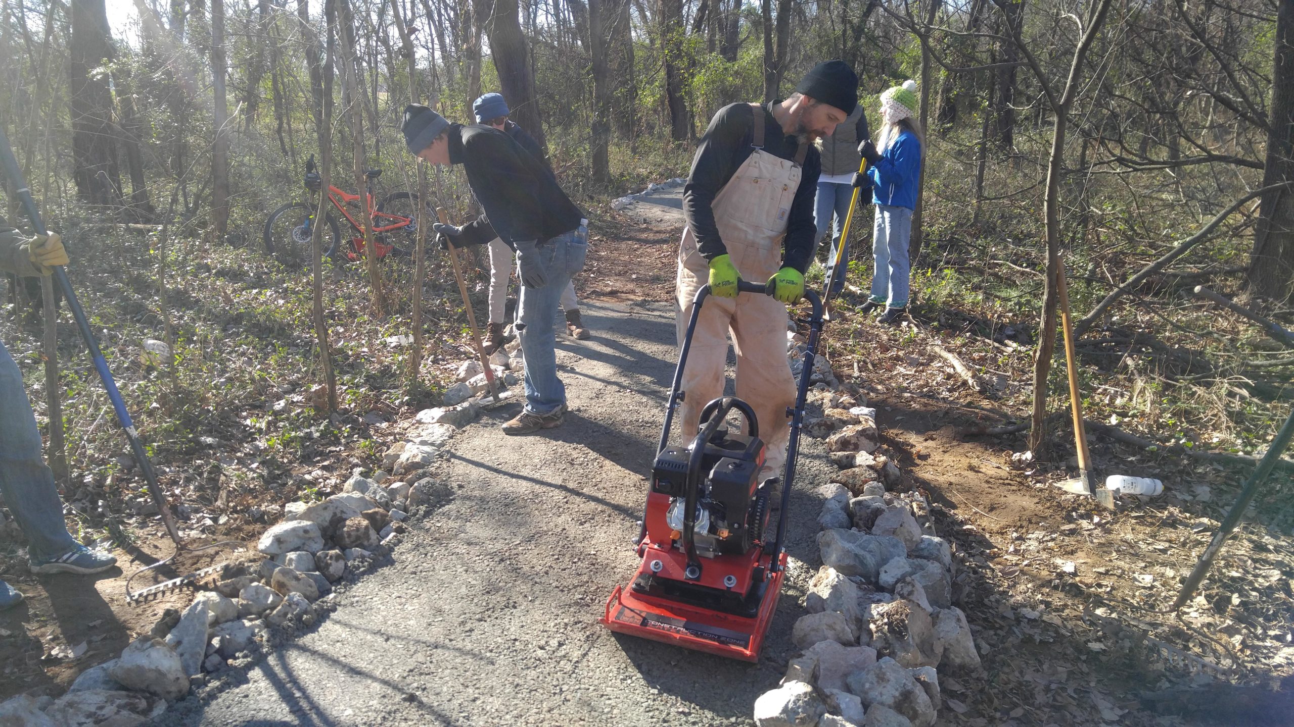 A group of volunteers working together to build a gravel path in a wooded area. Several individuals are using tools such as rakes and shovels, while one person operates a compacting machine. Rocks outline the path, and a bicycle is visible in the background. Ancarrow