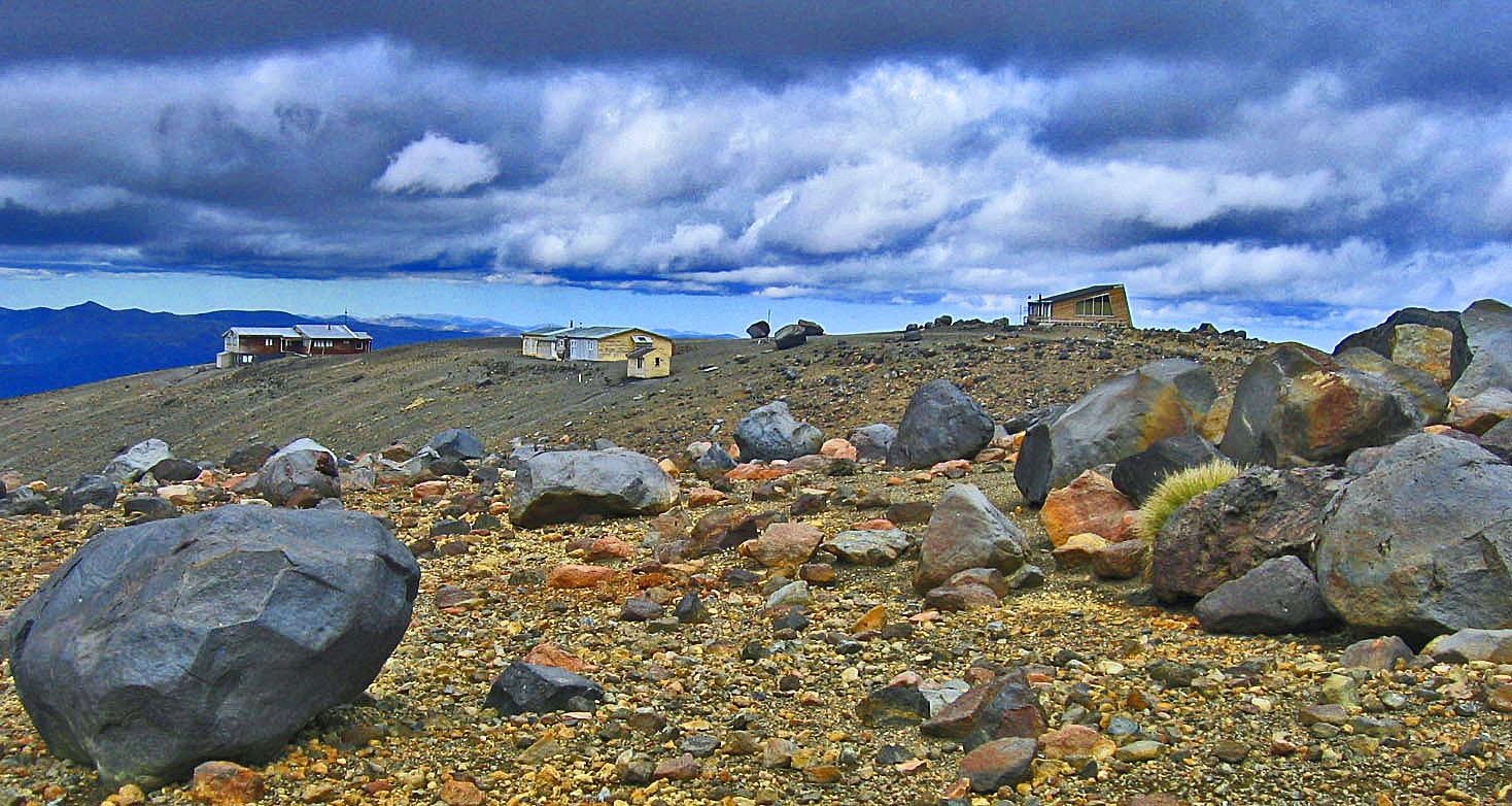 A rocky landscape with large stones in the foreground, leading to a gently sloping hill. In the background, there are several buildings with varying architectural styles, set against a dramatic sky filled with clouds. Mountains can be seen in the distance. Tukino Skifield Road mountain bike trail.