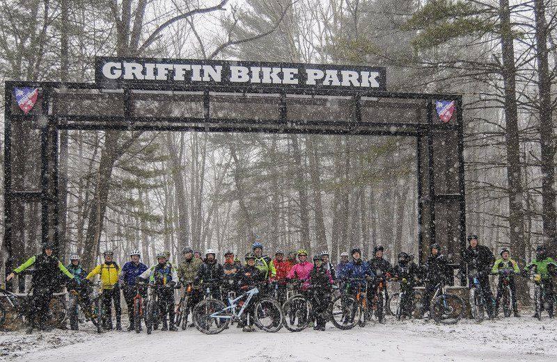 A group of cyclists stands in front of the Griffin Bike Park entrance, surrounded by tall trees and falling snow, showcasing a winter biking scene. Entrance Road mountain bike trail.