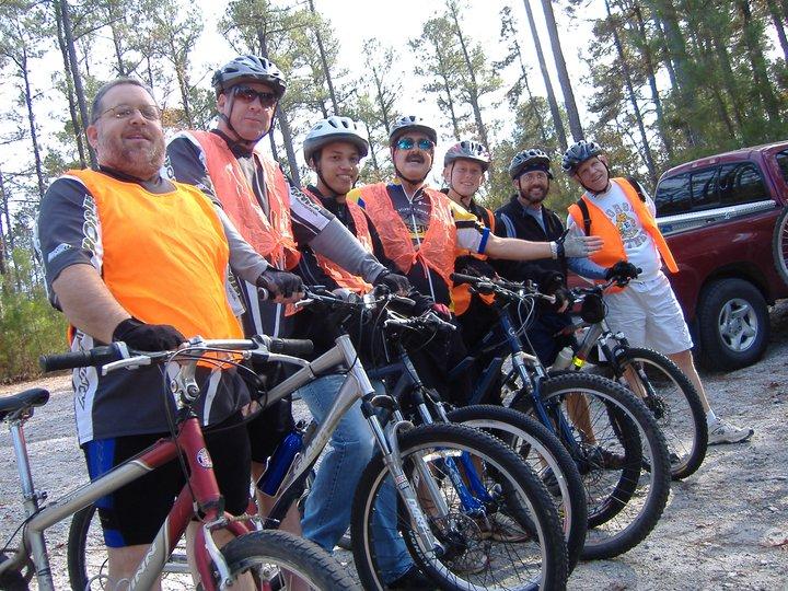 Specialized SWORKS Stumpjumper FSR: A group of seven people dressed in biking gear, including helmets and bright orange vests, stand next to their mountain bikes in a forested area. They are smiling and posing for the photo, with trees visible in the background and a parked truck on the right.