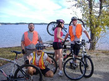 Specialized SWORKS Stumpjumper FSR: Four cyclists wearing bright orange vests are gathered by a lake. Two men and one woman stand beside their bicycles, while another man is squatting down, holding a bike tire. The scene is set against a backdrop of water and trees under a clear sky.