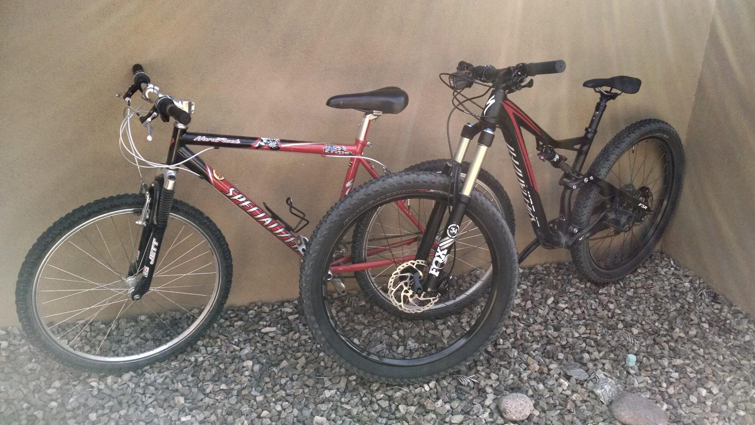 Specialized Stumpjumper FSR comp carbon 650B: Two bicycles resting against a wall, one red and silver mountain bike and one black mountain bike, both positioned on a gravel surface. The red bike features a noticeable spring front fork and traditional handlebars, while the black bike has a modern design with wider tires and a suspension system. Soft light rays illuminate the scene.