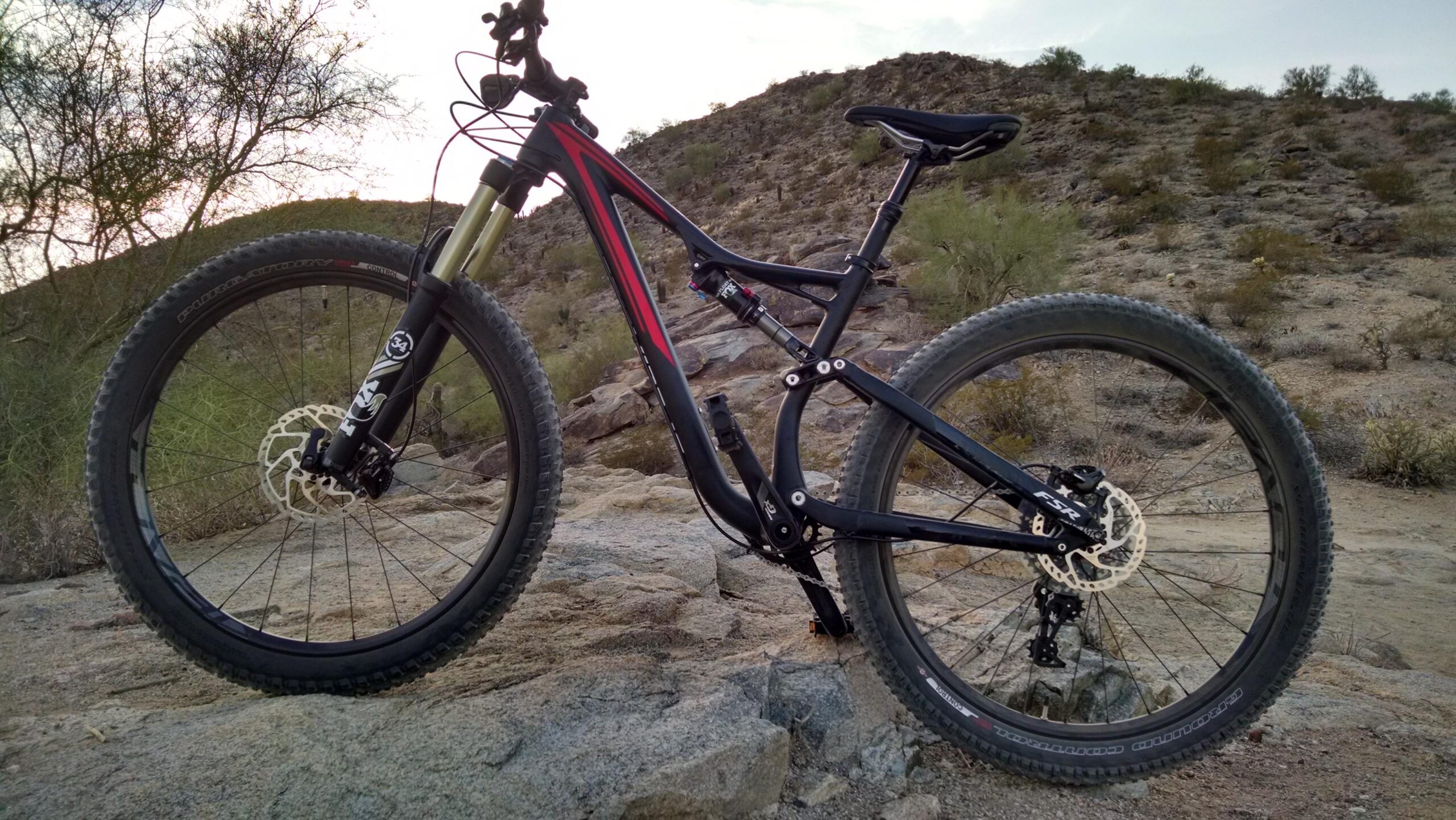 Specialized Stumpjumper FSR comp carbon 650B: A mountain bike is positioned on rocky terrain, featuring a black frame with red accents and thick, knobby tires. The background shows a desert landscape with hills and sparse vegetation, under a cloudy sky.