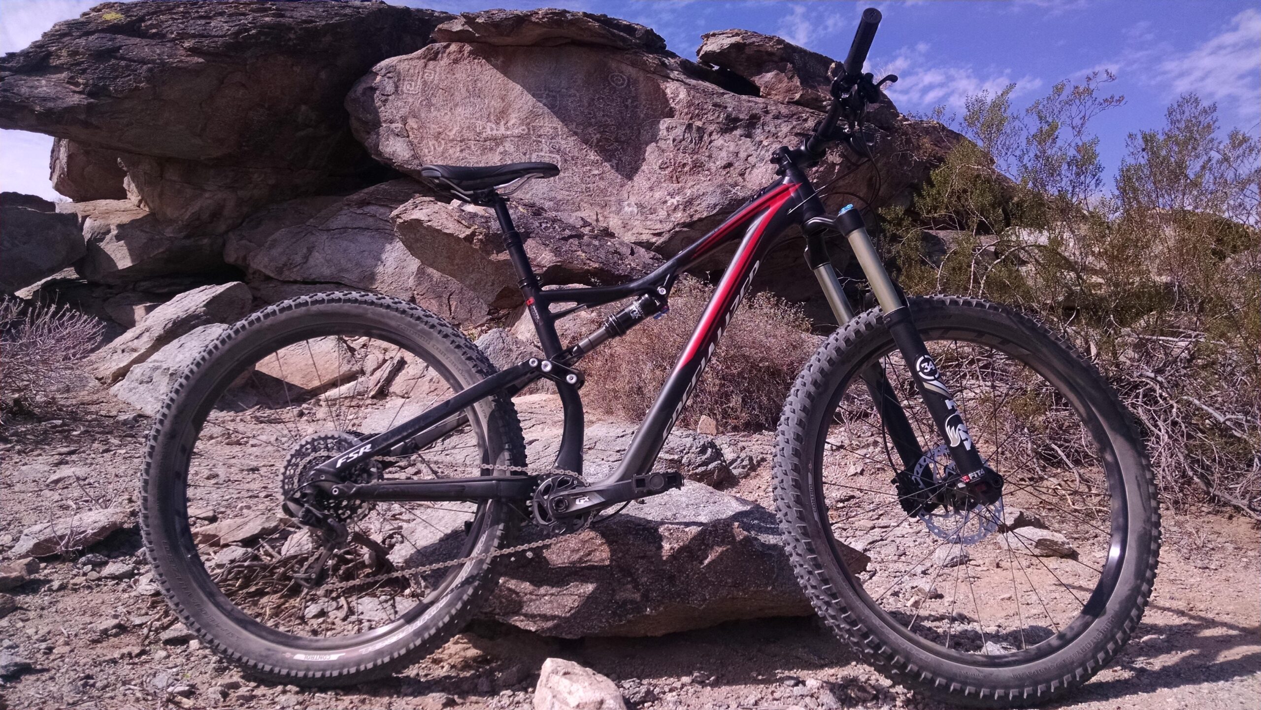 Specialized Stumpjumper FSR comp carbon 650B: A mountain bike positioned beside large rocky outcrops in a rugged, outdoor environment. The bike features a black and red frame, thick tires suitable for off-road terrain, and visible suspension components. The background includes dry shrubs and a clear blue sky.