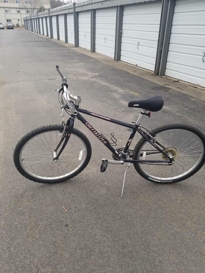 Specialized Rockhopper: A black mountain bike is parked on a paved surface, with storage units visible in the background. The bike features a prominent front shock, thick tires, and a standard seat. Its frame has a slight wear and tear, suggesting it has been used. The surrounding area is clear with no obstacles.