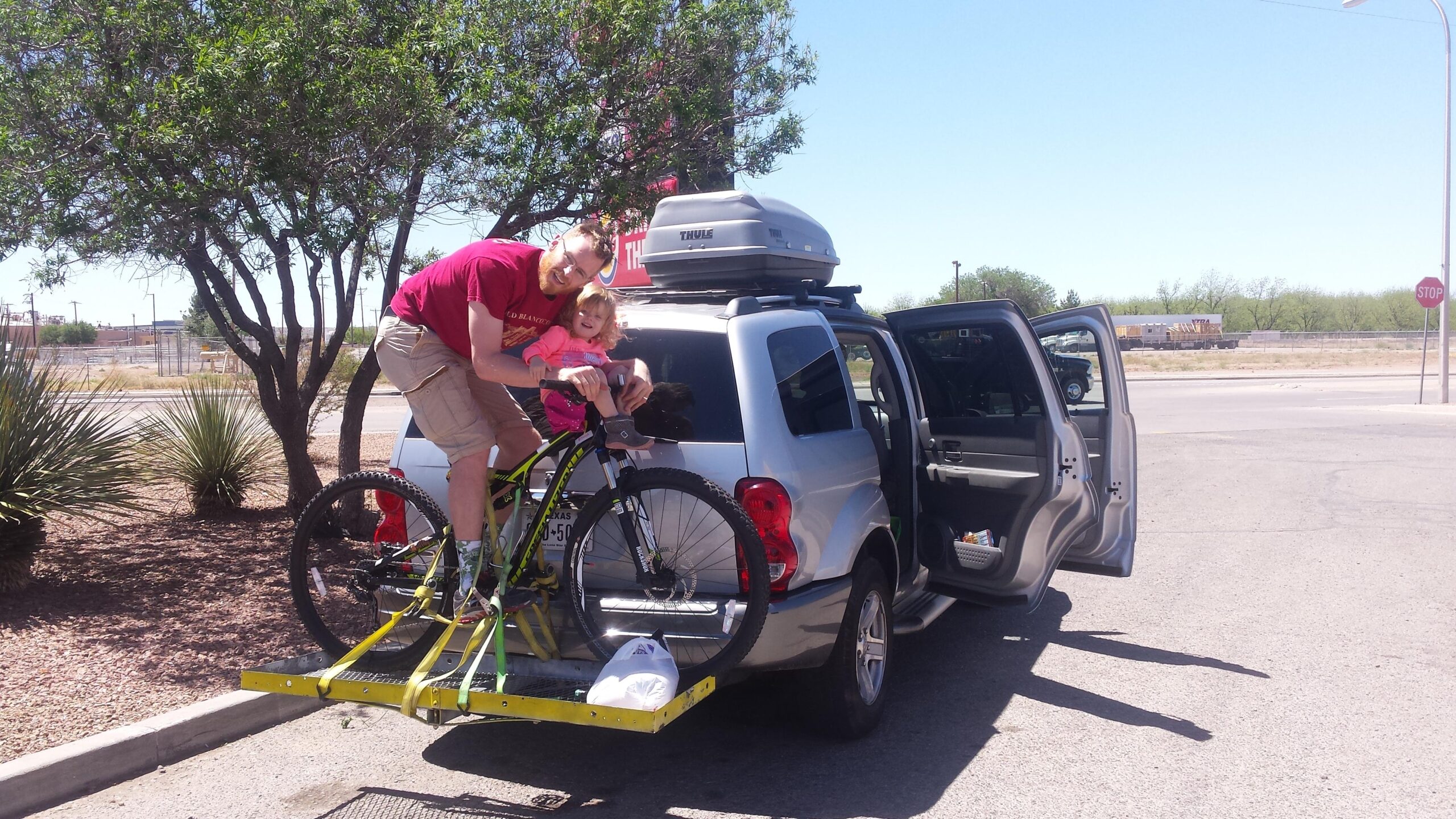Specialized Camber Comp 29: A man and a young child are enjoying time together at a parking lot, with the child seated on a bicycle attached to a car rack. The man is playfully posing next to the child, who is smiling. The car, a silver SUV, has its doors open, and a rooftop cargo box is visible. Surrounding them are a few small trees and shrubs, under a clear blue sky.