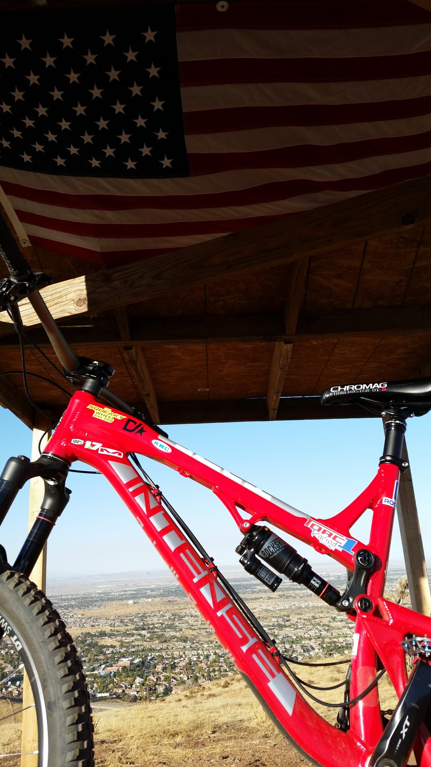 Intense Tracer 275: A close-up view of a red mountain bike frame with visible components and branding, positioned beneath a wooden structure. An American flag hangs above, and in the background, a panoramic view of a valley with houses and hills is visible under a clear blue sky.