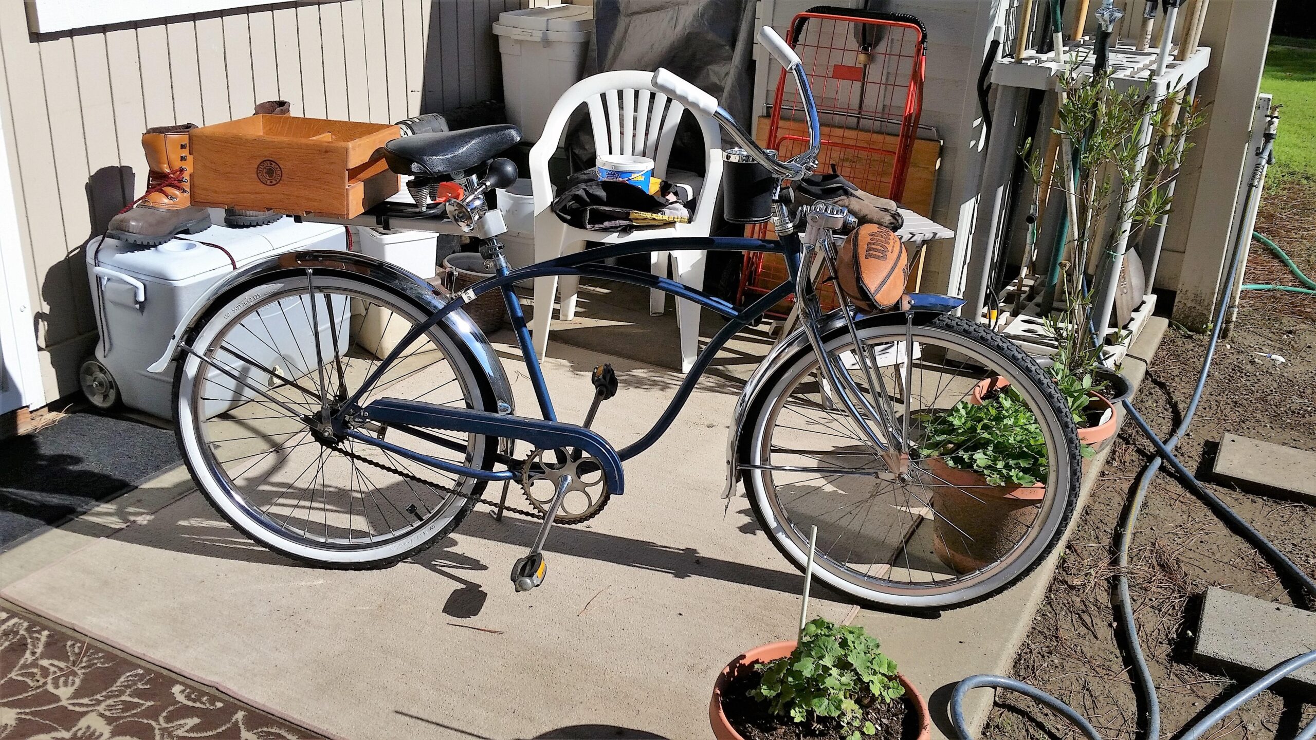 Schwinn Homegrown: A vintage bicycle with a blue frame and chrome accents is parked on a patio, surrounded by various items including a pair of work boots, a wooden box, and a cooler. In the background, there is a white folding chair and a collection of gardening tools. Potted plants are visible near the base of the bike, adding a touch of greenery to the scene.