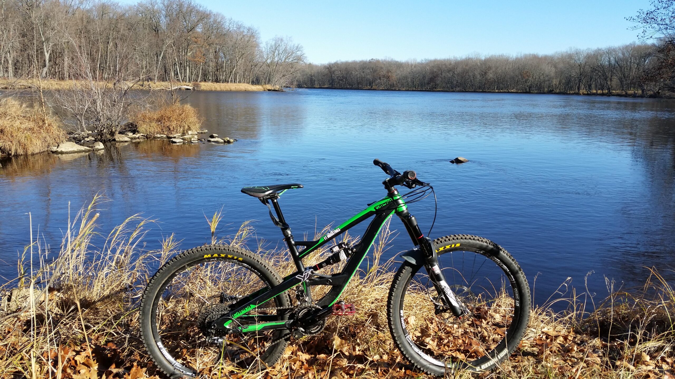 YT Capra: A mountain bike resting on the grass next to a calm river, surrounded by trees with bare branches and bushes in the background. The scene is bright, showcasing a clear blue sky and autumn foliage.